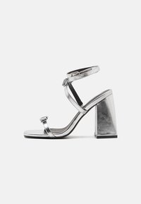 Metalické stříbrné sandály na vysokém podpatku s pásky kolem kotníku, čtvercovými blokovými podpatky a minimalistickým designem. Hladká textura a elegantní silueta.