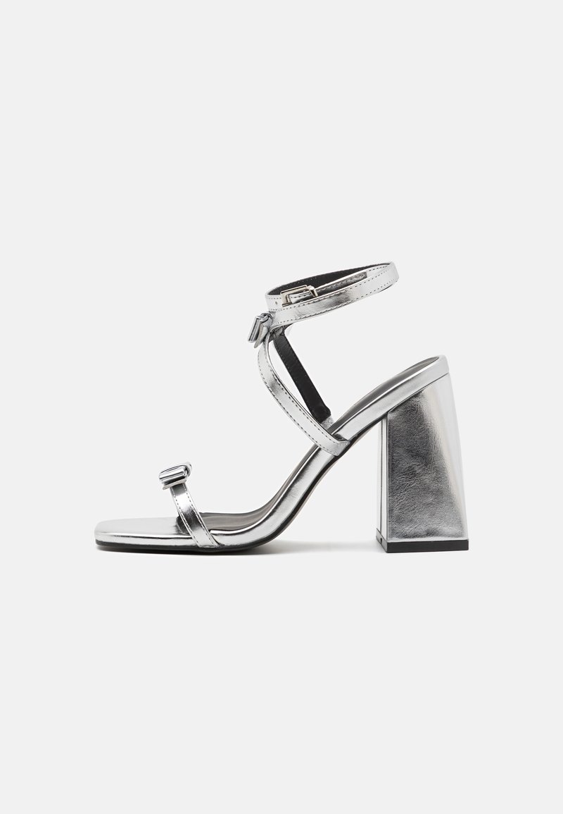 Metalické stříbrné sandály na vysokém podpatku s pásky kolem kotníku, čtvercovými blokovými podpatky a minimalistickým designem. Hladká textura a elegantní silueta.