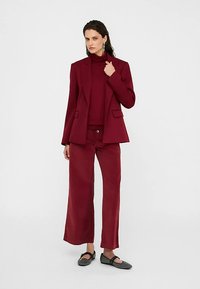 Vinrød blazer med hakkede revers, kombineret med matchende bukser med vide ben og en tætsiddende rullekrave. Sorte flade sko fuldender outfittet.