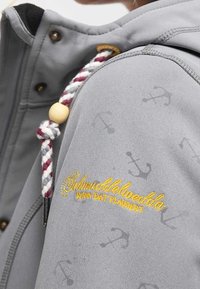 Detailní záběr na rukáv šedé bundy se subtilními vzory kotvy, žlutým vyšívaným textem a proplétanou šňůrkou v barvách vínové, bílé a šedé s dřevěnou korálkou.