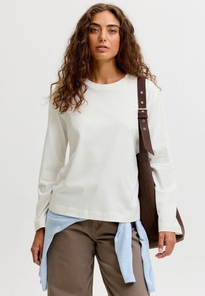 Femme aux cheveux bouclés bruns porte une chemise blanche à manches longues, un pantalon marron, un pull bleu clair noué à la taille et porte un sac à épaule marron.