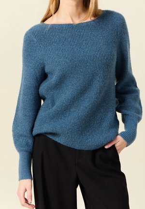 Kobieta w niebieskim swetrze o fakturze, z długim rękawem i czarnych spodniach, stojąca z jedną ręką w kieszeni na beżowym tle.