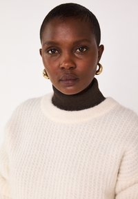 Maglione bianco lavorato a maglia con un motivo testurizzato e un ampio scollo a girocollo color crema, indossato sopra un dolcevita marrone scuro. Orecchini a cerchio dorati.