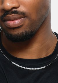 Zilveren draadketting met kleine, glanzende witte parels in een symmetrisch patroon, gedragen over een zwart t-shirt.