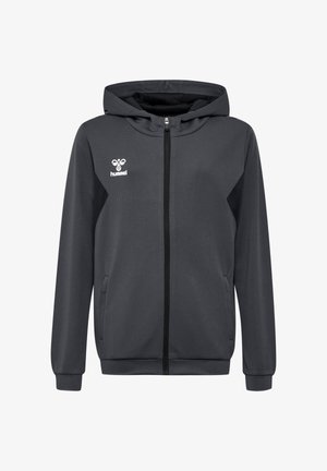 Ung kvinde står med den ene hånd i lommen iført sort lynlås-hættetrøje og sorte træningsbukser med chevronstriber på siderne.