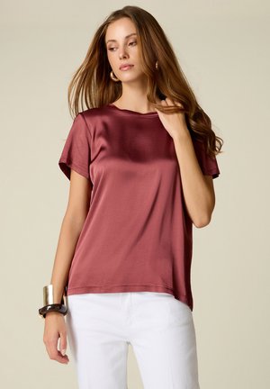 Kvinde med langt brunt hår iført bordeaux satin kortærmet top, hvide bukser, guldringe øreringe og chunky armbånd, kigger ned.
