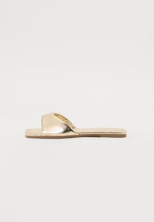 Sandale dorée à enfiler avec un design supérieur incurvé. Surface métallique lisse, semelle plate et style minimaliste sans quincaillerie supplémentaire.