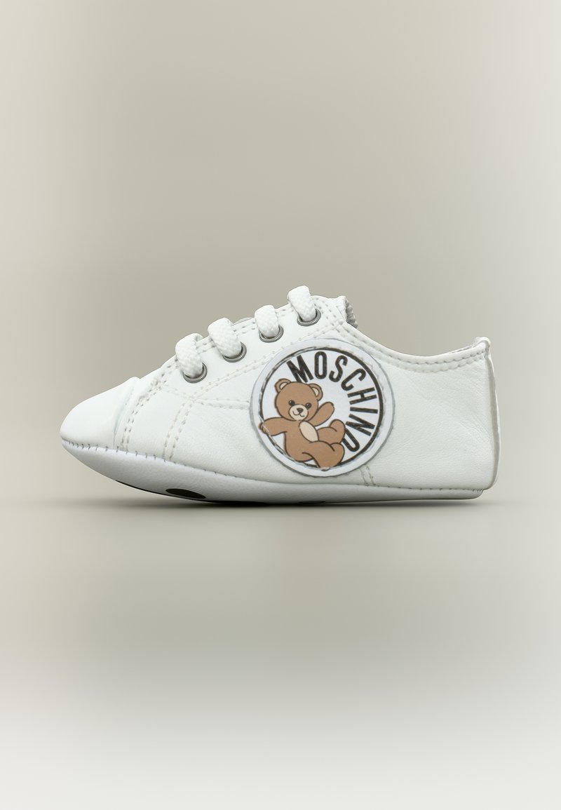 Λευκό παιδικό αθλητικό παπούτσι με λευκά κορδόνια και στρογγυλή επιγραφή στο πλάι που απεικονίζει μια αρκούδα και το κείμενο "MOSCHINO".