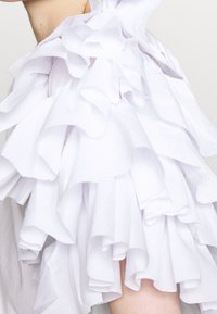 Valkoinen paita, jossa on kerroksellisesta kankaasta valmistettu röyhelöhiha. Pinta on pehmeä ja paita on ilmava ja runsas, korostaen röyhelöiden muotoja.