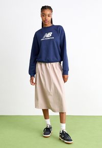 Navy collegepaita, jossa valkoinen "New Balance" -logo, beige midi-hame, mustat lenkkarit valkoisilla ja vaaleanruskeilla yksityiskohdilla, vihreää taustaa vasten.