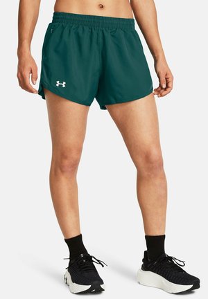 Kobieta ubrana w zielone sportowe spodenki Under Armour, czarne skarpetki i czarne buty do biegania, stojąca na prostym jasnym tle.