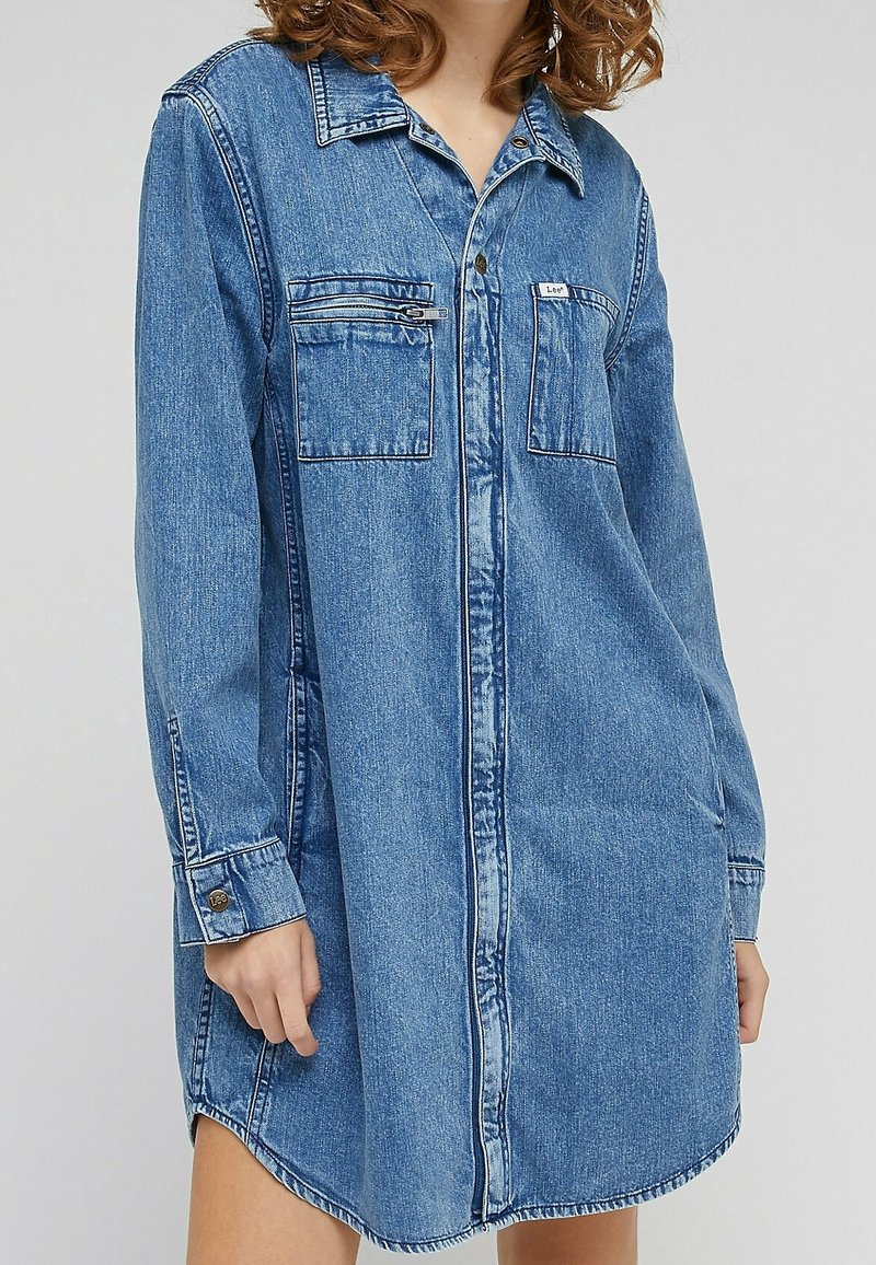 Robe-chemise en denim à manches longues, boutonnée devant, avec deux poches poitrine et un col, portée par une personne aux cheveux bouclés.