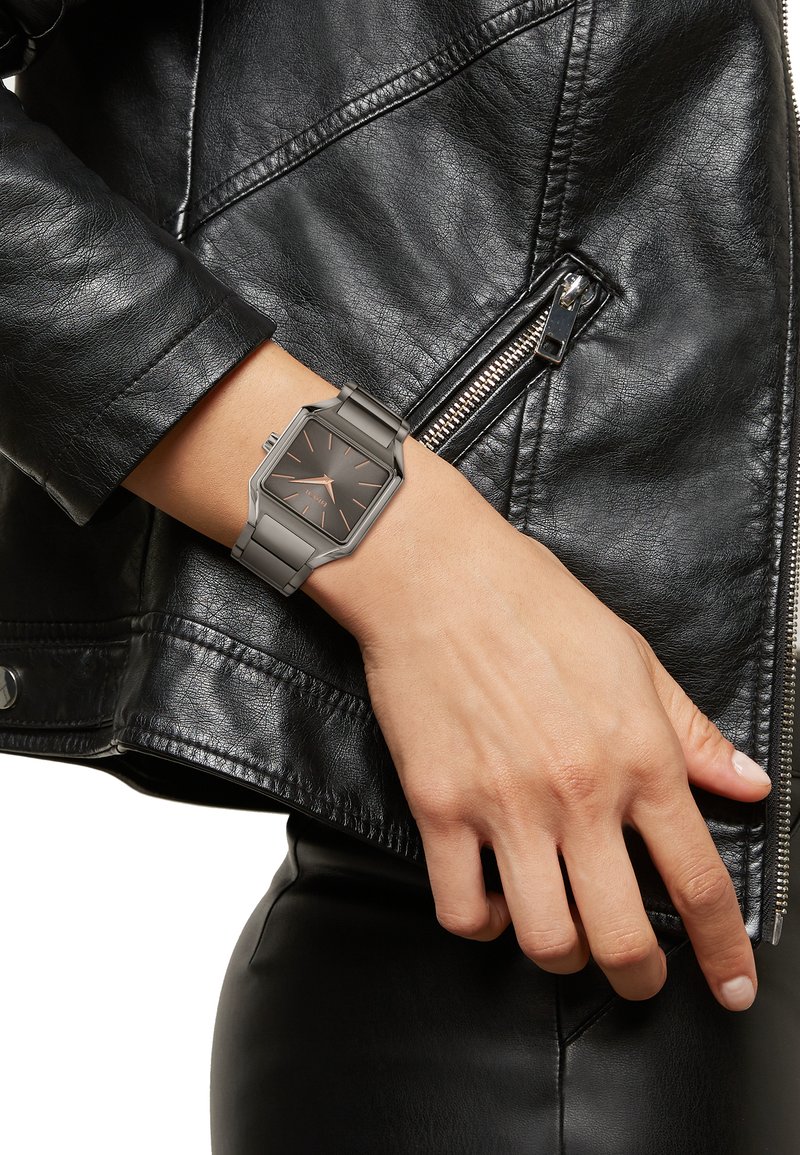 Montre en métal gris avec un cadran rectangulaire et un fond sombre, dotée d'accents en or rose. Portée sur un poignet à côté d'une veste en cuir noire.