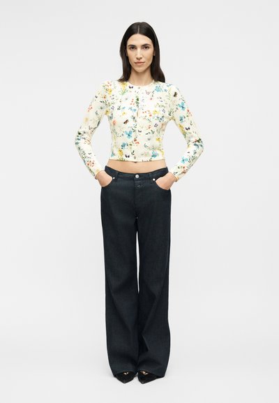 Γυναίκα στέκεται με τα χέρια στις τσέπες φορώντας εφαρμοστό φλοράλ πουλόβερ με κουμπιά και μαύρο παντελόνι φαρδιάς γραμμής ψηλόμεσο, μπροστά σε λευκό φόντο.