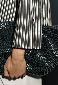 Black woven clutch bag held in a hand, with a black and white striped shirt sleeve partially visible. The clutch features a textured surface.