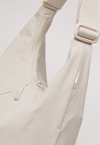 Beige Umhängetasche aus einem glatten, leichten Stoff, mit Reißverschluss und minimalistischen Design. Verfügt über einen kontrastierenden Tragegurt und ein kleines Logo-Detail.