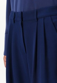 Pantaloni plissettati blu navy realizzati in tessuto morbido, con vita alta, chiusure a doppio gancio e tasche laterali.