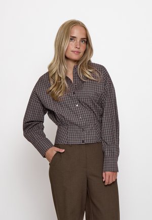 Donna bionda con capelli sciolti indossa una camicia marrone a quadri con bottoni e pantaloni marroni, in piedi con una mano in tasca su sfondo bianco.