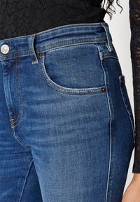 Džínové kalhoty v tmavě modré barvě s hladkou texturou, které mají klasický design, pět kapes a akcenty v oranžovém prošití.