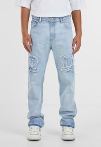 Jeans en denim bleu clair avec des patchs déchirés, ornés de grandes lettres audacieuses, d'une coupe décontractée et d'une finition à lavage moyen.