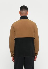 Pull en polaire marron et noir, avec un col haut et des manches longues. Le design comprend un motif en color-block avec une texture douce.