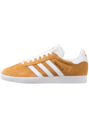 Zapatilla de ante en color beige con rayas blancas de Adidas, cordones blancos, texto con la marca "GAZELLE" y suela de goma blanca texturizada.
