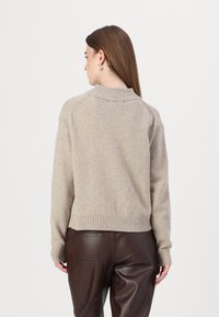 Pulover bej din tricot cu guler înalt, manșete și tiv cu nervuri. Partea din spate are o croială lejeră și sleeve raglan. Purtați-l peste pantaloni maro cu textură.