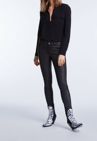 Femme portant un chemisier noir à boutons, un pantalon en cuir noir et des bottines à motif peau de serpent noir et blanc, debout sur un fond neutre.