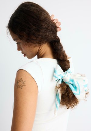 Ženska z rjavimi kodrastimi lasmi, spletenimi in zvezanimi z belo in modro črtasto pentljo, oblečena v belo brezrokavnik, ki kaže tetovažo sonca na zgornjem delu roke.