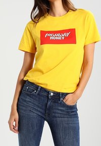 Žluté bavlněné tričko s červeným obdélníkovým potiskem "HIGHWAY HONEY" v bílé barvě. V kombinaci s tmavě modrými džíny, středním střihem.