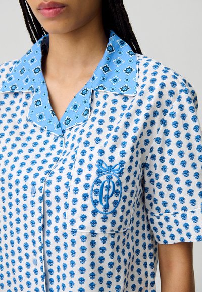 Camicia a maniche corte blu e bianca con un motivo floreale, chiusura frontale a bottoni e un'emblema ricamato sulla tasca sinistra.