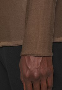 Ruskea pitkähihainen paita pehmeästä, teksturoidusta kankaasta, istuvalla leikkauksella ja tikatulla helmalla, yhdistetty mustiin, vartalonmyötäisiin housuihin.