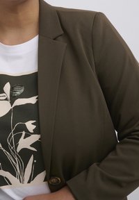 Blazer vert olive avec une coupe structurée, des revers crantés et une fermeture à boutons, porté sur un t-shirt blanc avec un imprimé botanique.