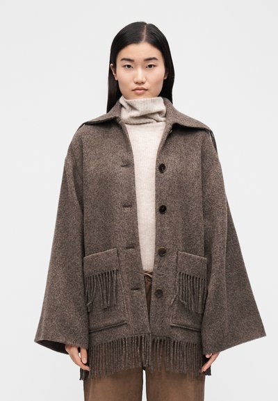 Barna gyapjúkeverék kabát gallérral, gombos elülső résszel, nagy zsebekkel és rojtos díszítéssel az alján. Világos bézs garbó fölött viselve.