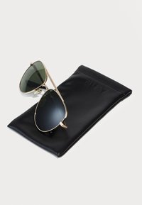 Occhiali da sole aviator dorati con lenti scure, poggiati su un astuccio in pelle nera con impressa la scritta "QUAY". Montatura lucida con naselli regolabili.