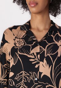 Sort bluse med et stort beige blomsterprint, med krave og knaplukning foran. Lavet af glat, let stof.