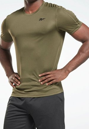 Mężczyzna ubrany w oliwkowo-zieloną sportową koszulkę Reebok z krótkim rękawem i czarne spodenki, stojący z rękami na biodrach na białym tle.