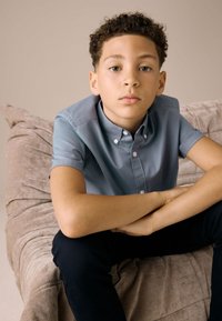 Sininen lyhythihainen kauluspaita, jonka napit ovat edessä, pojan päällä, joka istuu teksturoidulla beige-sohvalla, tummansiniset housut, neutraali tausta.