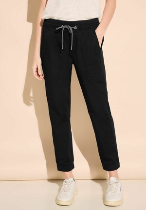 Joggers neri con vita elasticizzata e coulisse, tasche laterali, gambe affusolate e orli a costine, abbinati a sneakers bianche su uno sfondo neutro.