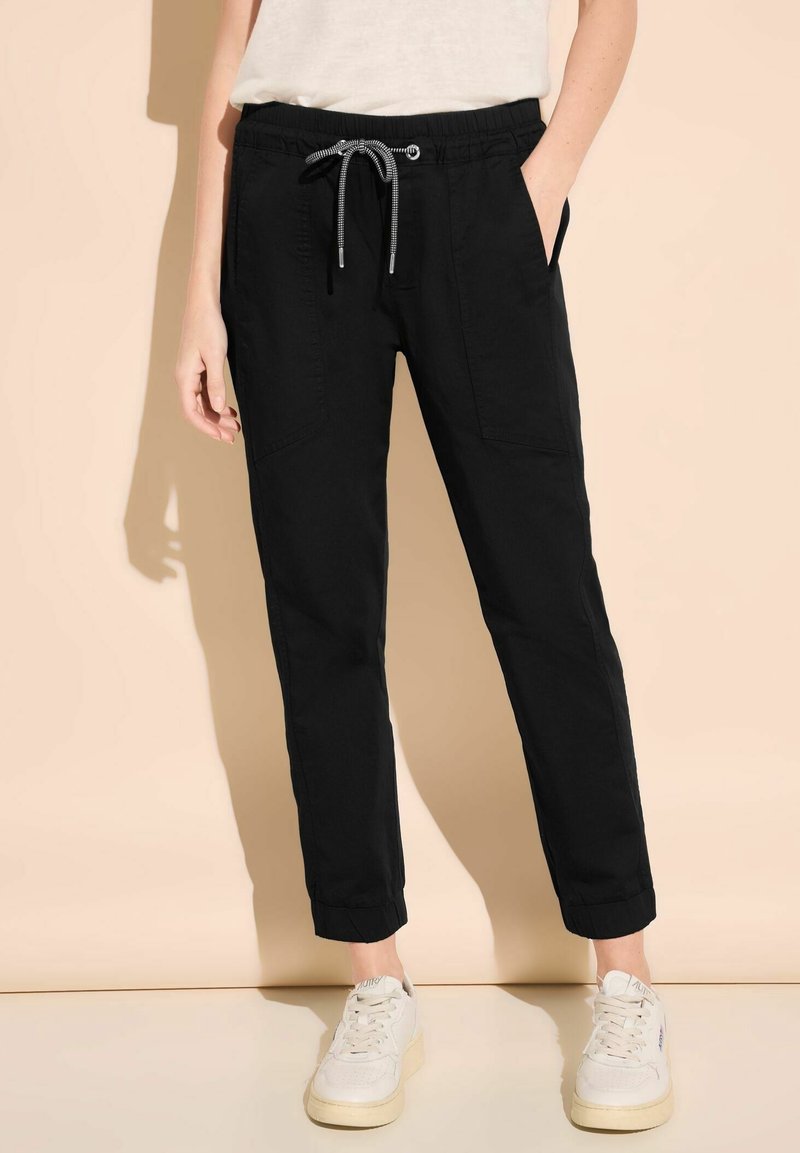 Svarta joggers med elastisk midja och dragsnöre, sidfickor, avsmalnande ben och uppvikta fållar, i kombination med vita sneakers mot en neutral bakgrund.