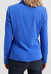 Blå skräddarsydd kavaj med markerade axlar, slätt tyg, slits bak och figursydd design. Bärs över mörk denim.