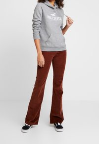 Felpa grigia con cappuccio e testo bianco, abbinata a pantaloni a zampa d'elefante in velluto a coste marroni e sneakers nere. Abbigliamento casual e rilassato.
