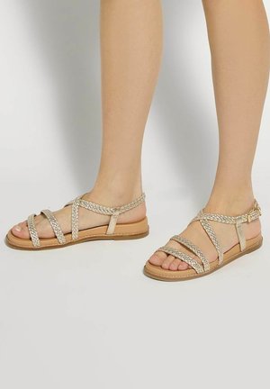Legs wearing flat tan sandals with braided straps and ankle buckles, standing on a light surface.