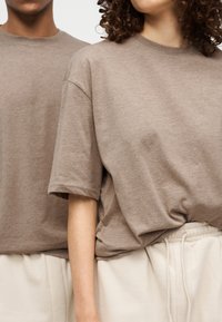 Δύο άτομα με μπεζ μπλουζάκια και ανοιχτό μπεζ παντελόνια, στέκονται πλάι-πλάι, με ορατό τον κορμό και το χέρι.