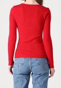 Haut rouge à manches longues côtelé avec un col en U, coupe ajustée et texture lisse, associé à un jean en denim bleu clair légèrement délavé.