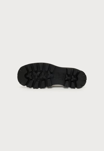 Semelle de chaussure noire avec des motifs de rainures profondes conçus pour l’adhérence et la durabilité, présentée sur un fond clair uni.
