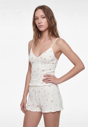 Jeune femme aux longs cheveux bruns portant un caraco côtelé blanc fleuri et un short assorti, posant avec la main sur la hanche sur un fond uni.