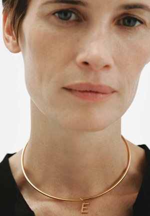 Primer plano de una persona que lleva un collar tipo choker liso de oro con un pequeño charm de la letra "E" en oro, vestida con una blusa negra.