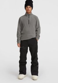 Grijze fleece pullover met kwart-rits en zwarte Gore-Tex broek. Een zwarte beanie en witte sneeuwlaarzen maken de outfit compleet.