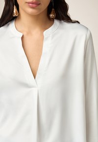 Blusa blanca con escote en V y cuello mao, que presenta una textura suave y una caída favorecedora. Pendientes de gota dorados complementan el conjunto.